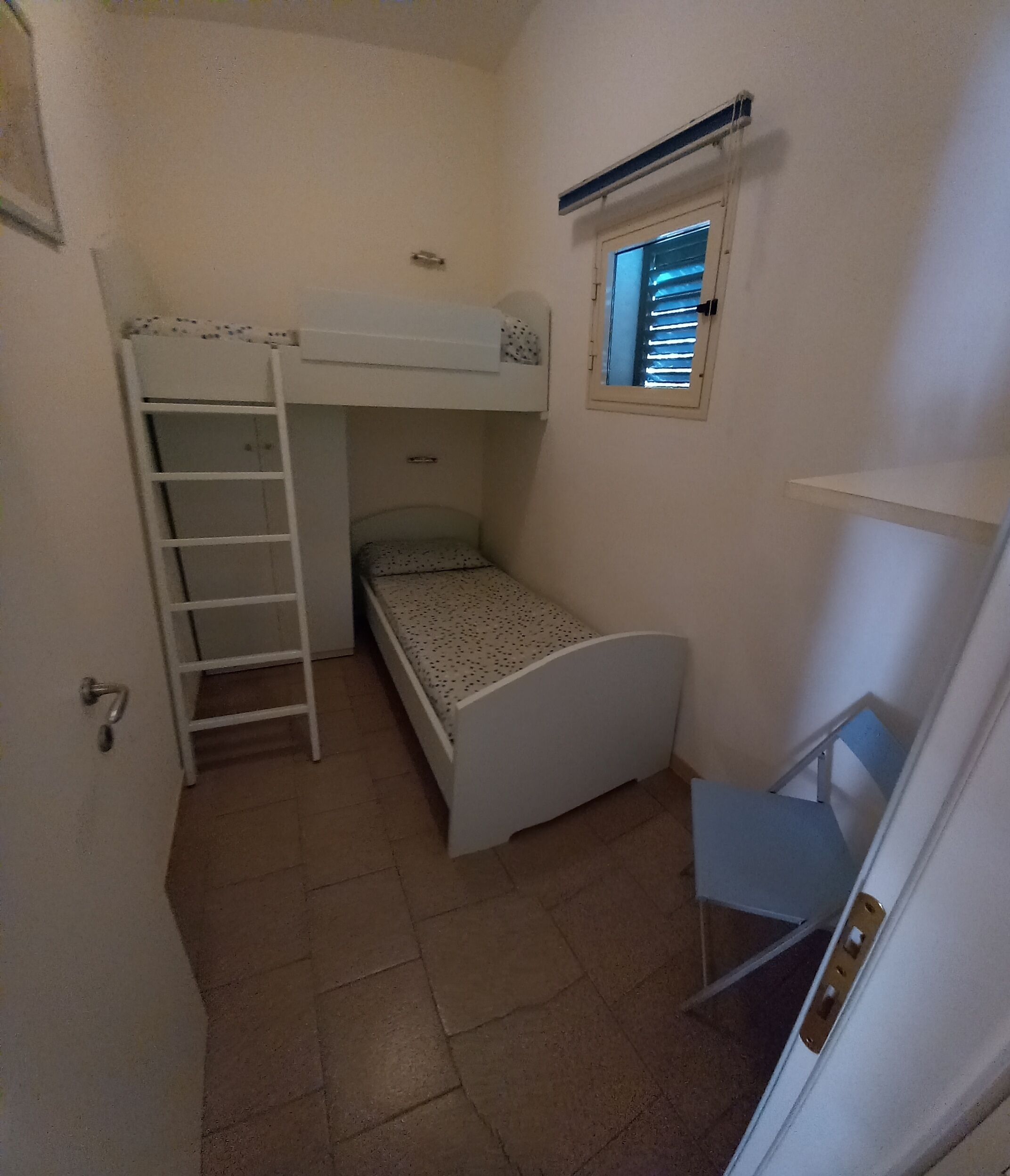 Ein kleiner, schmaler Raum mit einem Etagenbett, einem Einzelbett, einer Stehleiter, einem Fenster und einer Tür.
