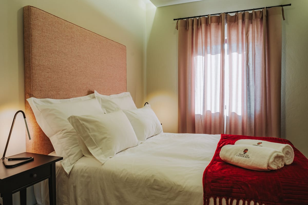 A neatly made bed with white linens and a red blanket, a bedside table with a lamp, and a window with curtains.