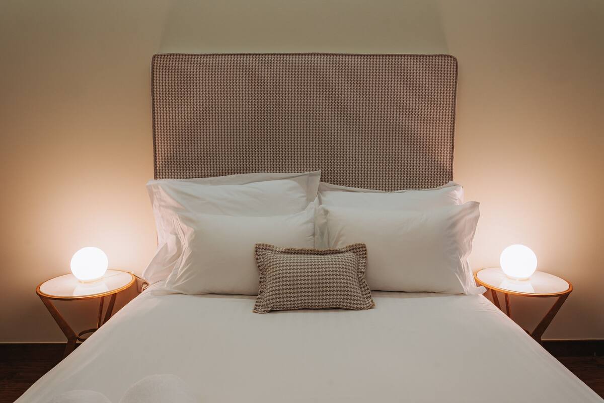 A neatly made bed with white linens and a brown headboard. There are two bedside tables with round wooden bases and a single round light on each.