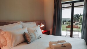 Una cama bien hecha con sábanas blancas y dos toallas con un logotipo, una lámpara de noche y vistas a un espacio exterior con bancos y vegetación.