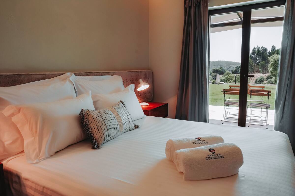 A neatly made bed with white linens and two towels with a logo, a bedside lamp, and a view of an outdoor area with benches and greenery.