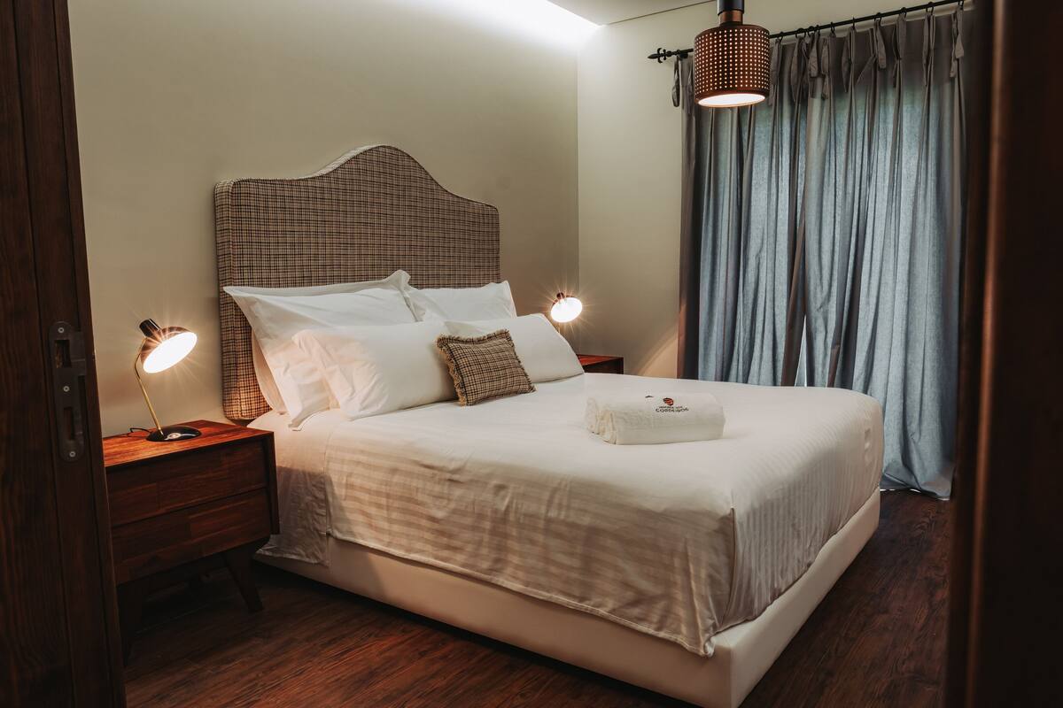 A neatly made bed with a checkered headboard, two bedside lamps, and a window with curtains.