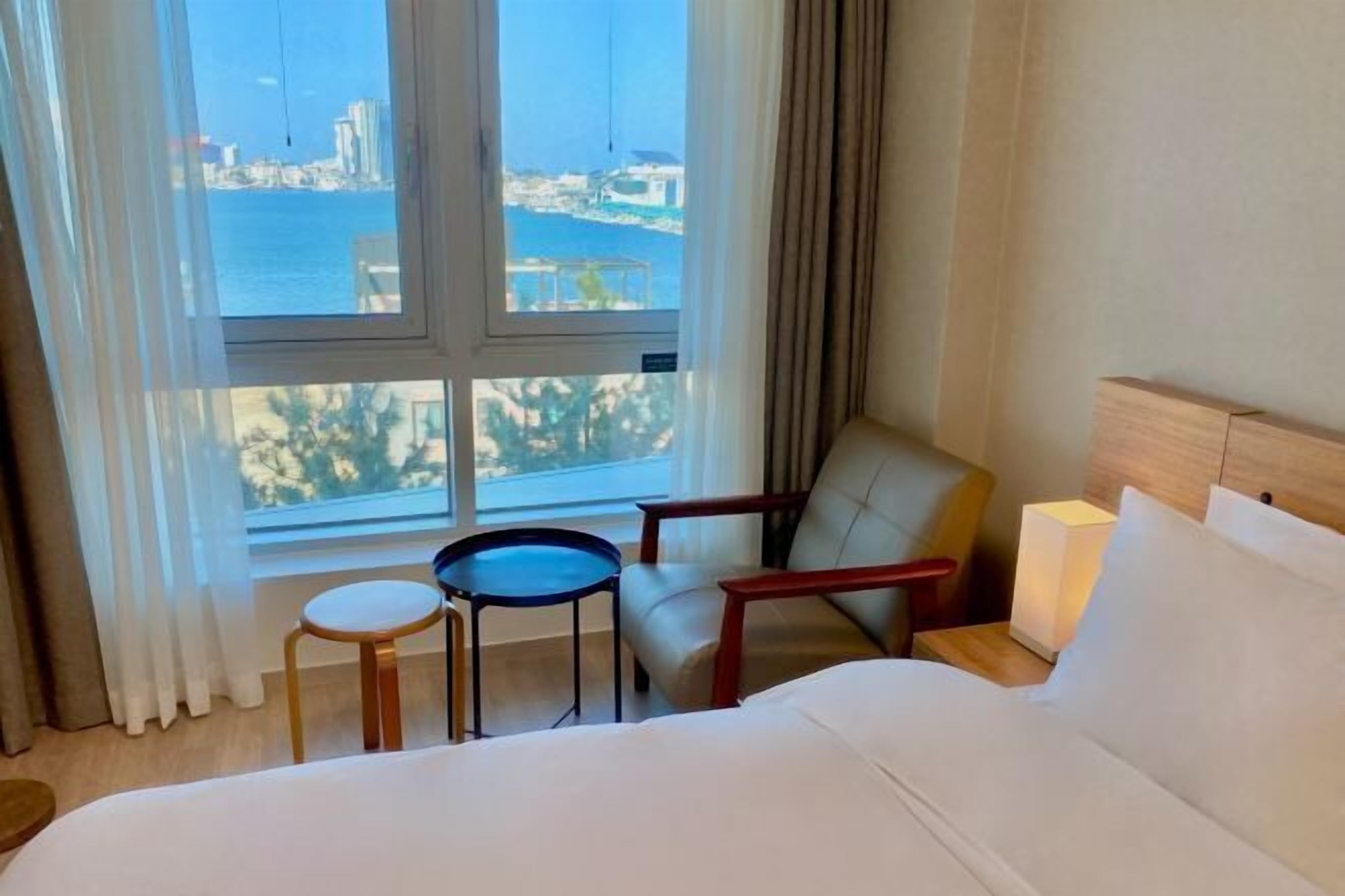 Ein Hotelzimmer mit Bett, einem kleinen Tisch, einem Stuhl und Blick auf das Meer und Gebäude.