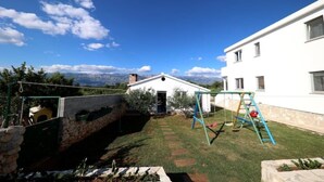A courtyard with a swing set, a stone wall, and a white building with a mountain view in the background. - Summer House Amon (Razanac)