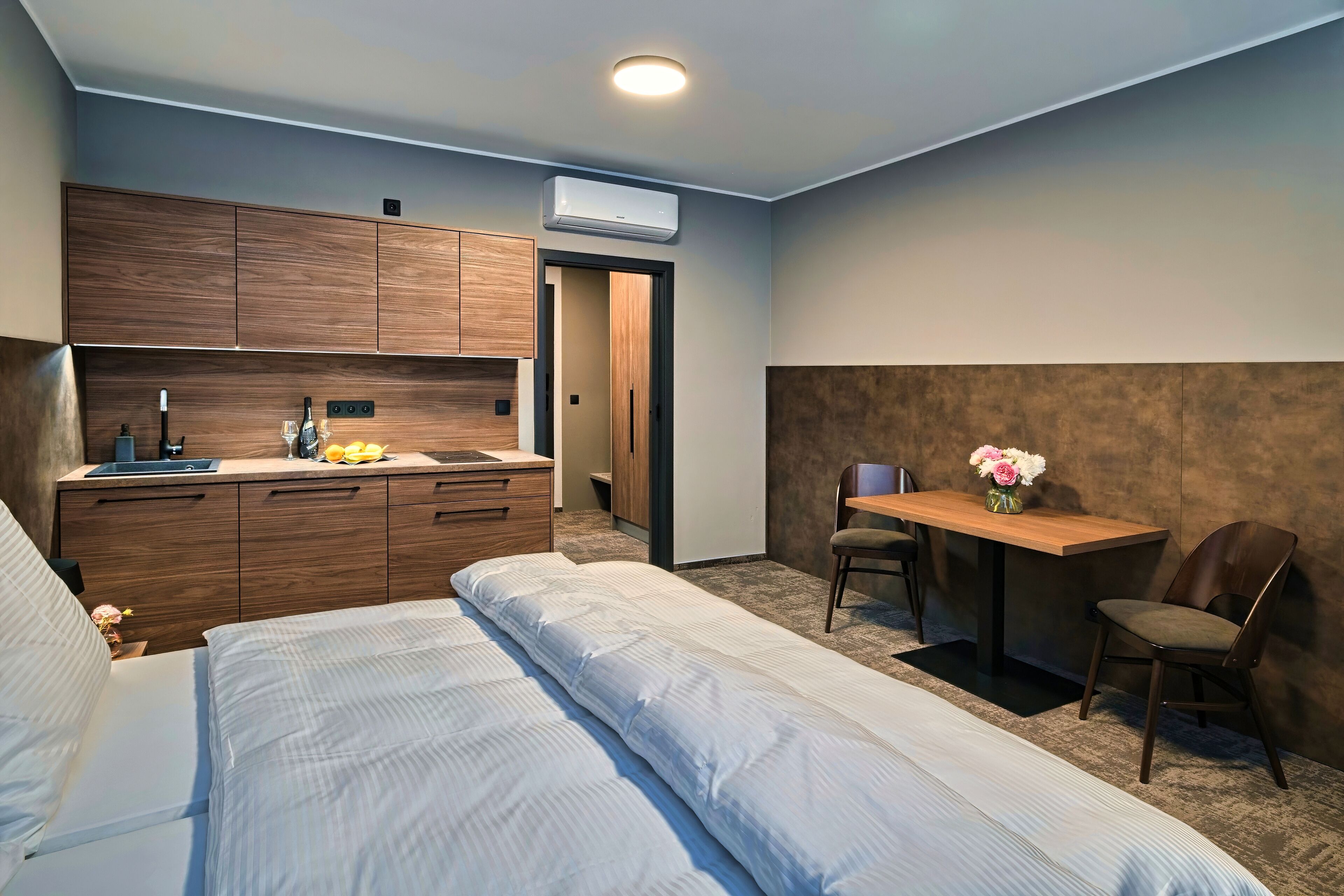 Habitación de hotel compacta con cama, una kitchenette pequeña, un escritorio con silla y aire acondicionado instalado en la pared.