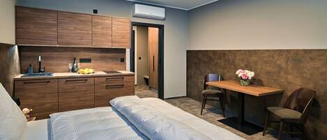 Habitación de hotel compacta con cama, una kitchenette pequeña, un escritorio con silla y aire acondicionado instalado en la pared.