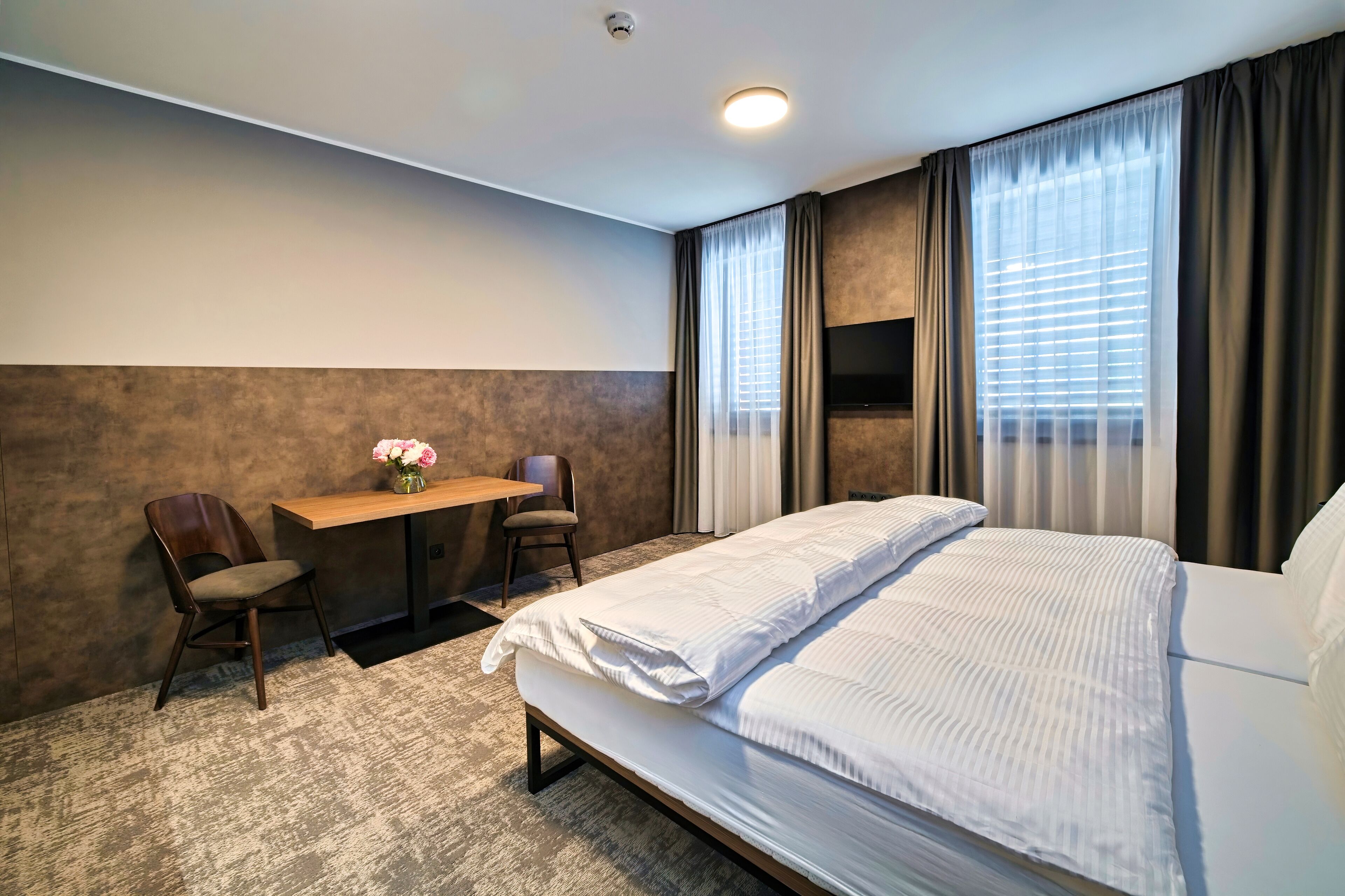 Een hotelkamer met een bed, een bureau, twee stoelen, een kleine tafel met bloemen en een televisie aan de muur.