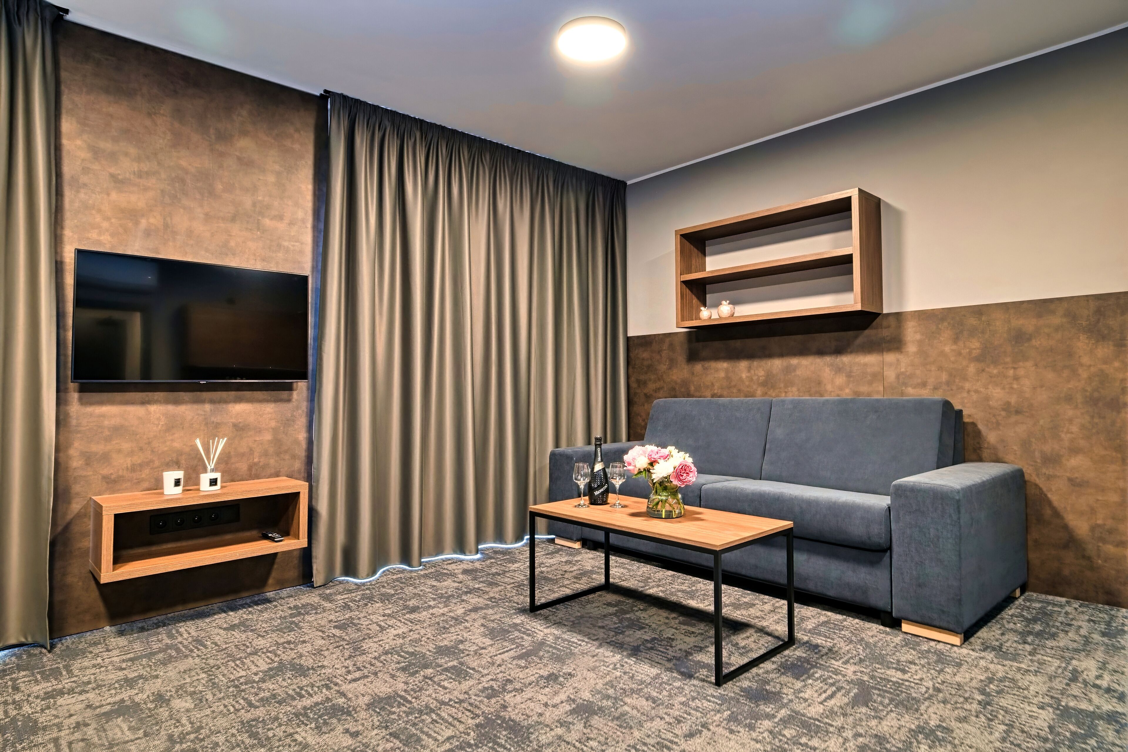 A modern living room with a flat-screen TV mounted on the wall, a grey sofa, a wooden coffee table with a vase of flowers, and a small shelf with decorative items.