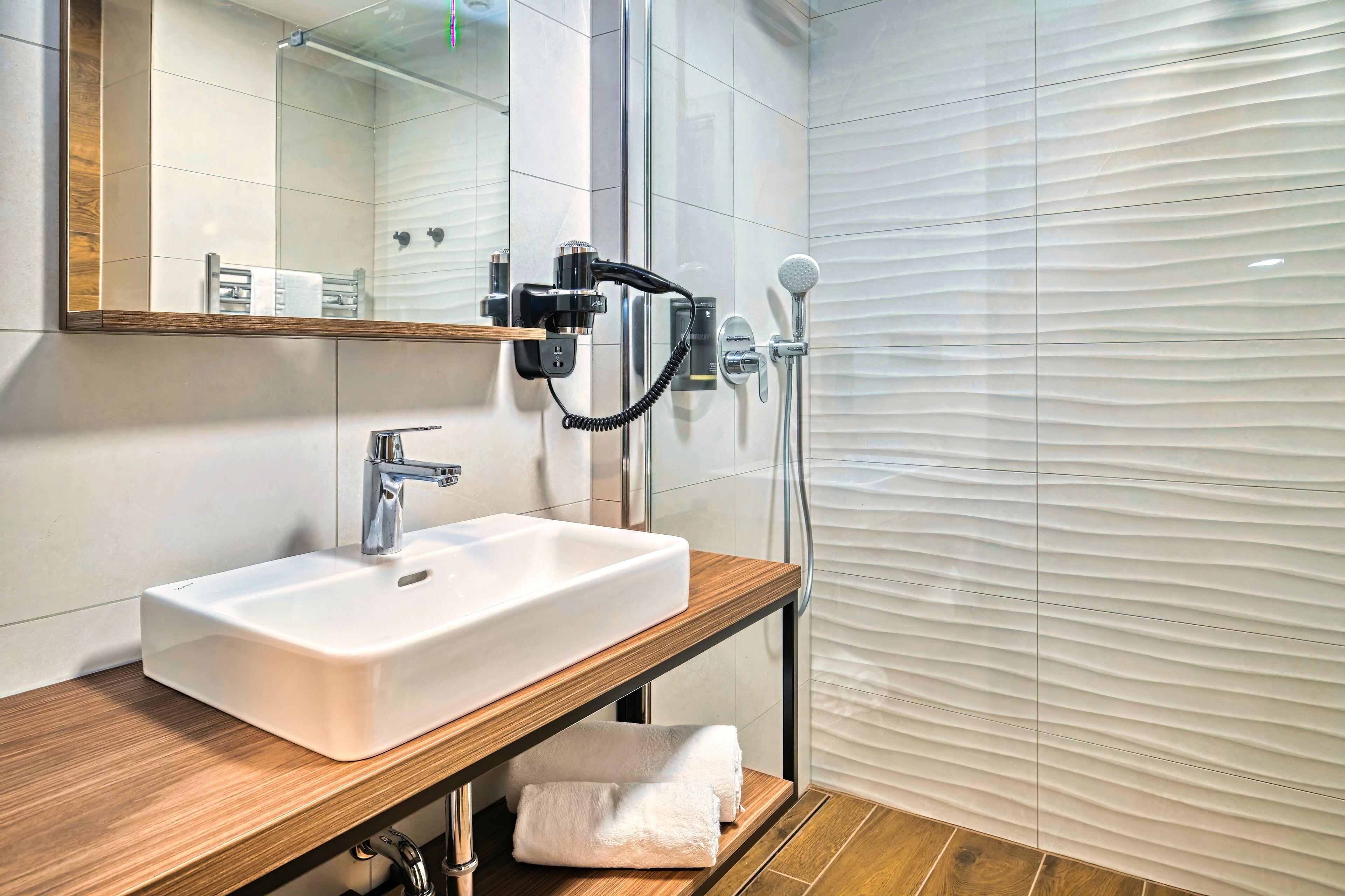 Une salle de bain moderne avec un lavabo blanc, un meuble-lavabo en bois, une cabine de douche en verre et un porte-serviettes.