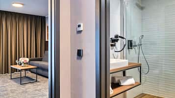 A modern hotel room with a sofa, a small table with a vase of flowers, a shower area with a handheld showerhead, and a control panel on the wall.