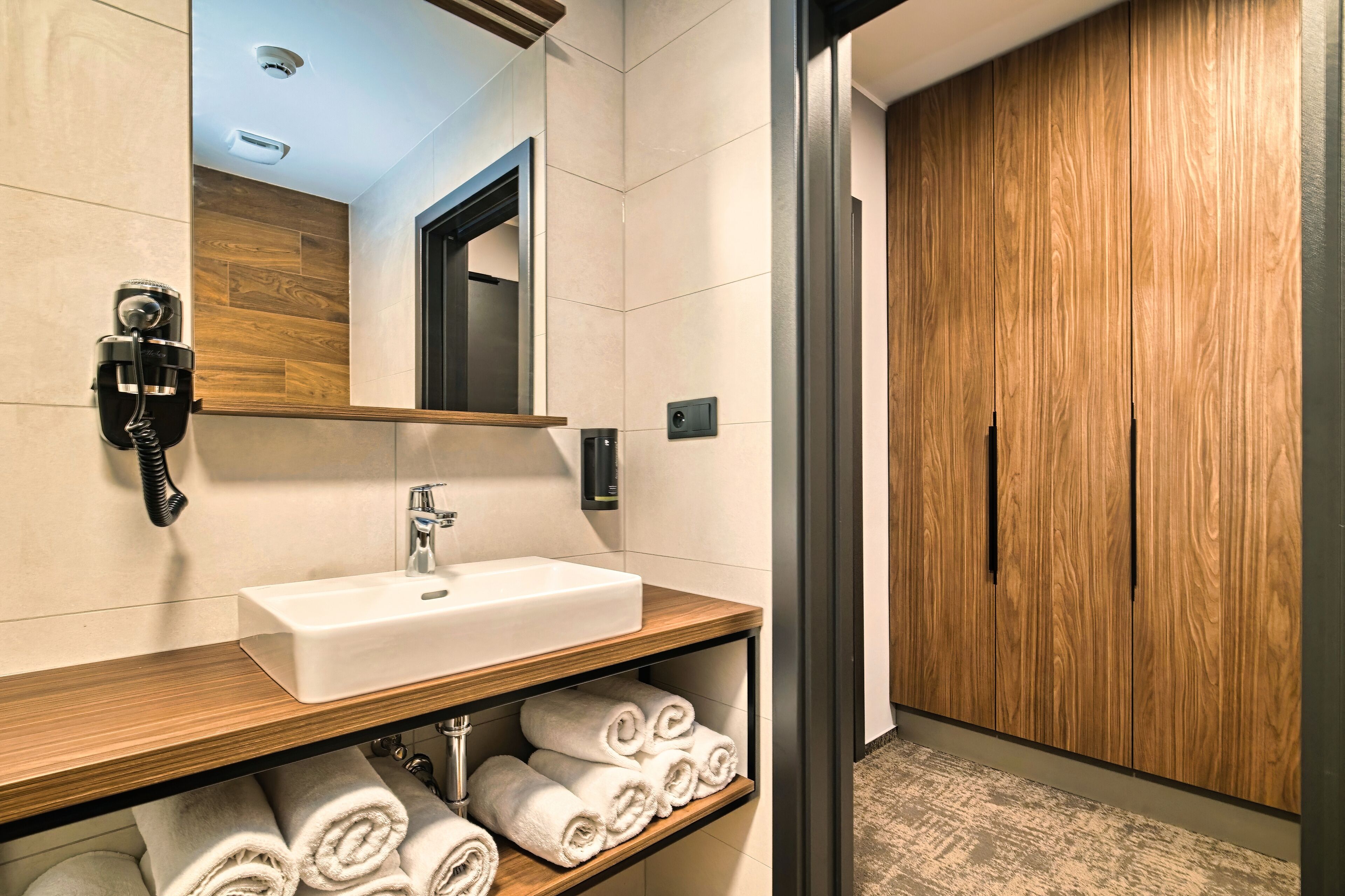 Un baño moderno con lavamanos blanco, tocador de madera y un espejo. Hay toallas en el estante y un armario de madera al fondo.