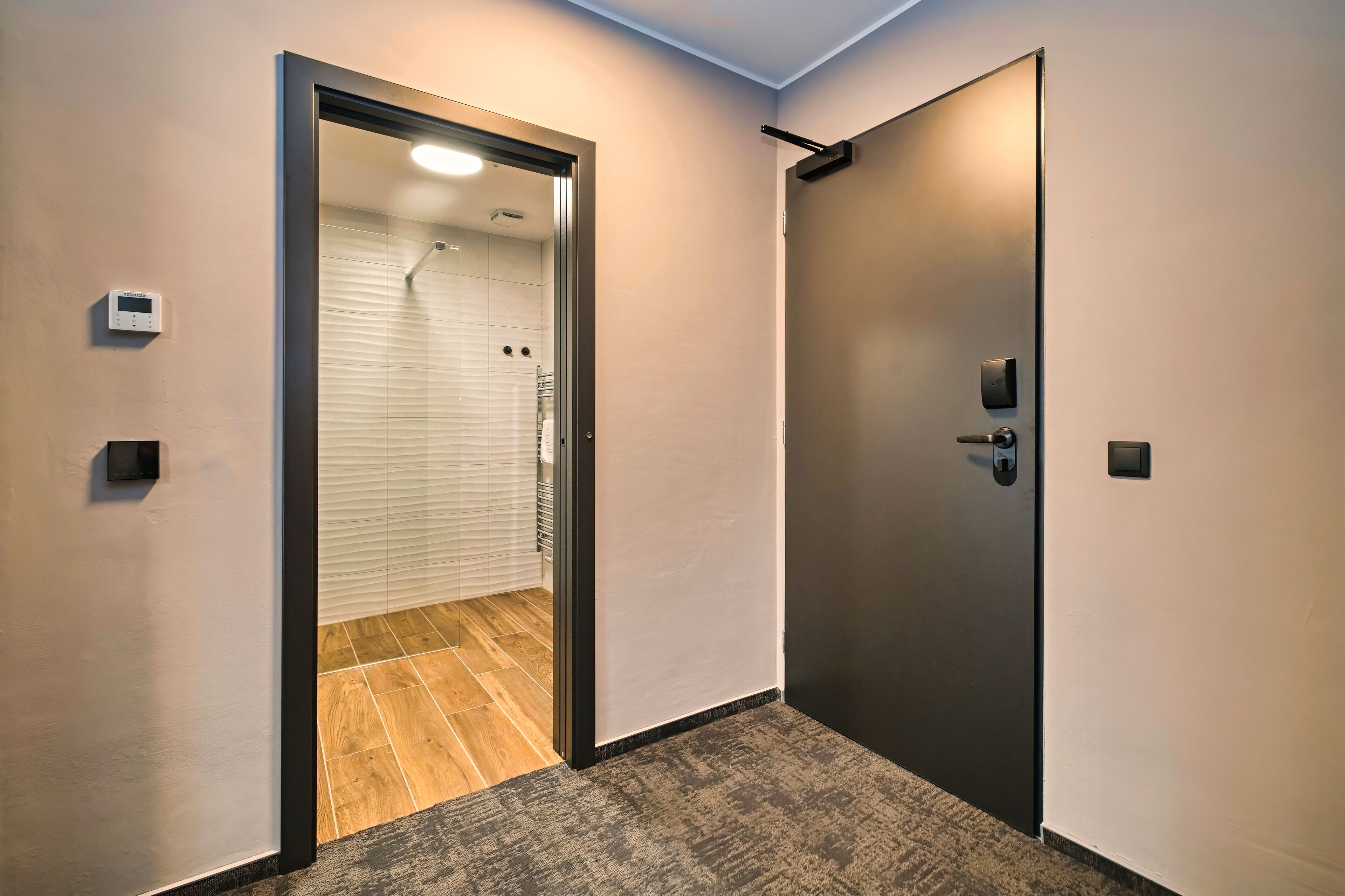 A modern hallway with a door leading to a bathroom, a thermostat on the wall, and a textured carpet.