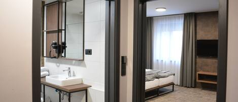 Una habitación de hotel moderna con baño que incluye un lavabo, un espejo y un tendedero para toallas. La habitación tiene una cama, un televisor y una ventana con cortinas.