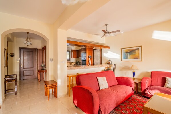 A living room with a red sofa, a wooden coffee table, and a kitchen area in the background. - Villa Pontus - stunning views & privacy in beautiful garden with pool & hot tub (Kouklia)