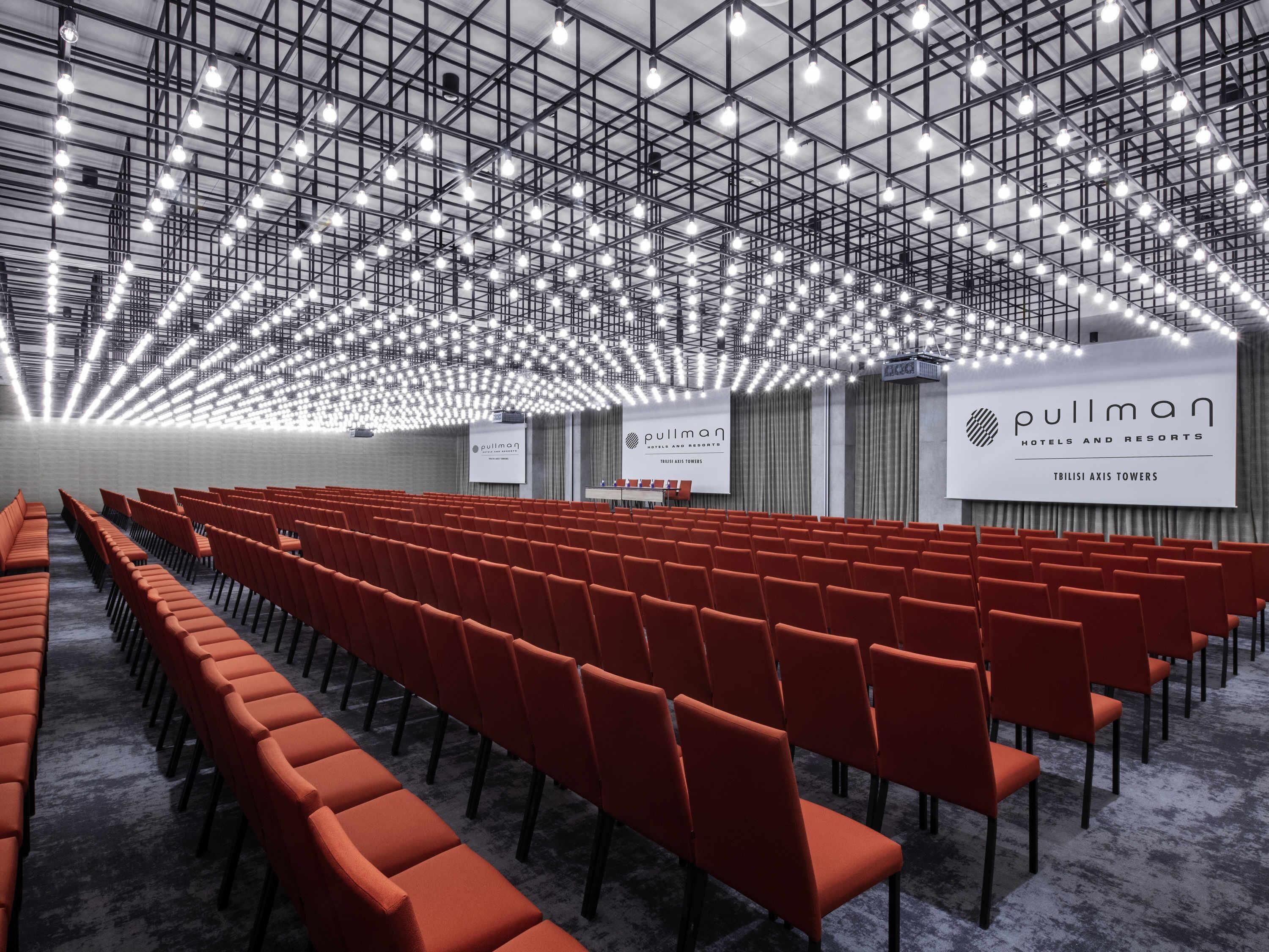 A conference room with rows of orange chairs, a high ceiling with hanging lights, and banners for Pullman Hotels and Resorts.