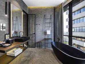 Une salle de bain moderne avec un grand miroir, deux vasques, une baignoire noire et une vue sur le paysage urbain grâce à de grandes fenêtres.