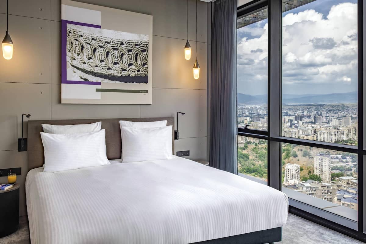 A modern hotel room with a large bed, a city view, and a framed artwork on the wall.