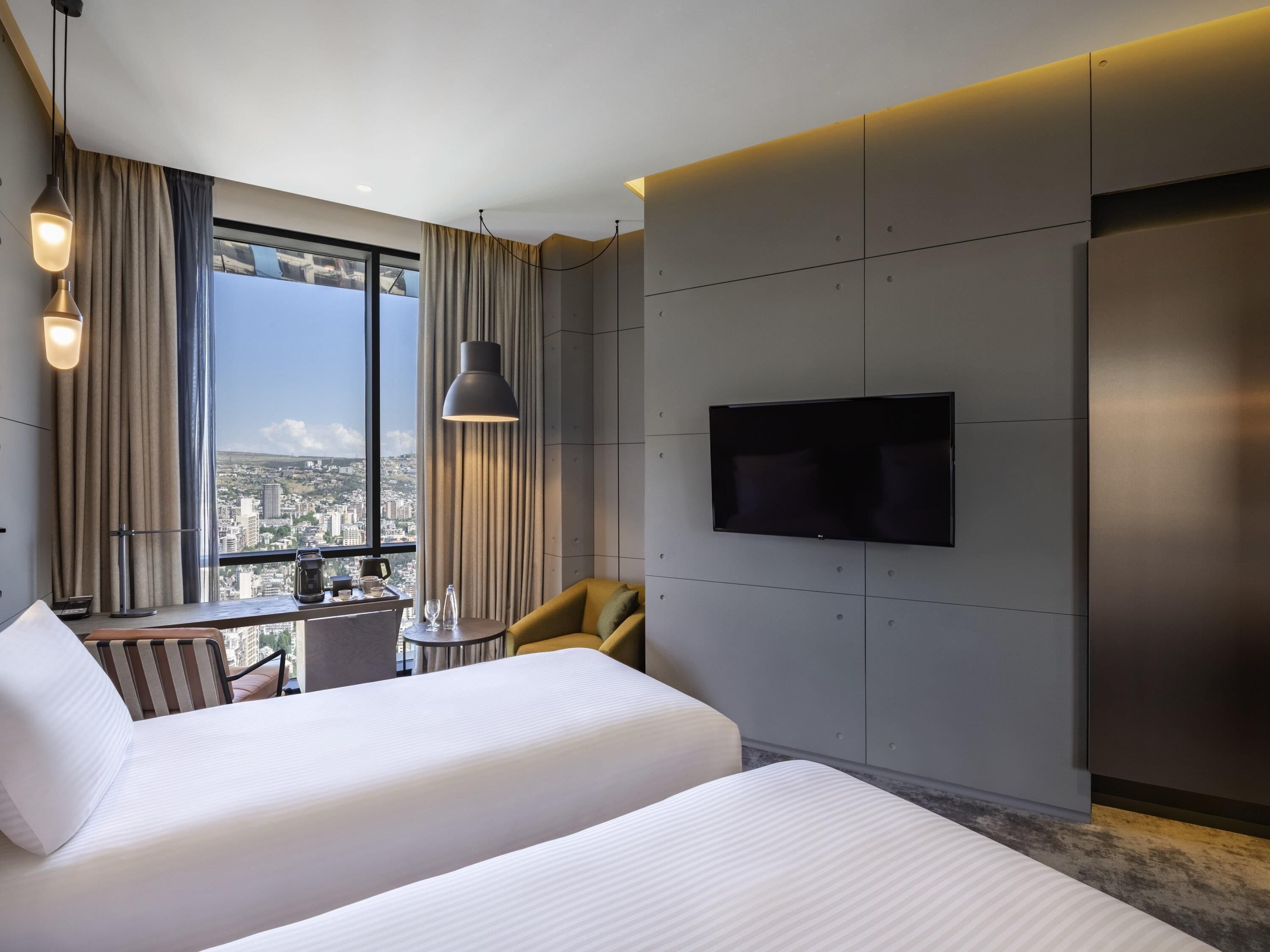Une chambre d’hôtel dotée d’une grande fenêtre, d’un téléviseur à écran plat fixé au mur et de deux lits avec du linge de lit blanc.