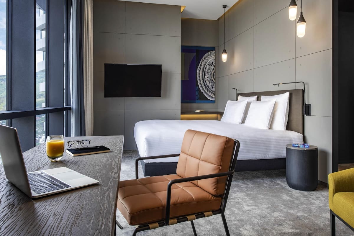 A modern hotel room with a bed, a desk with a laptop, a chair, and a TV on the wall.
