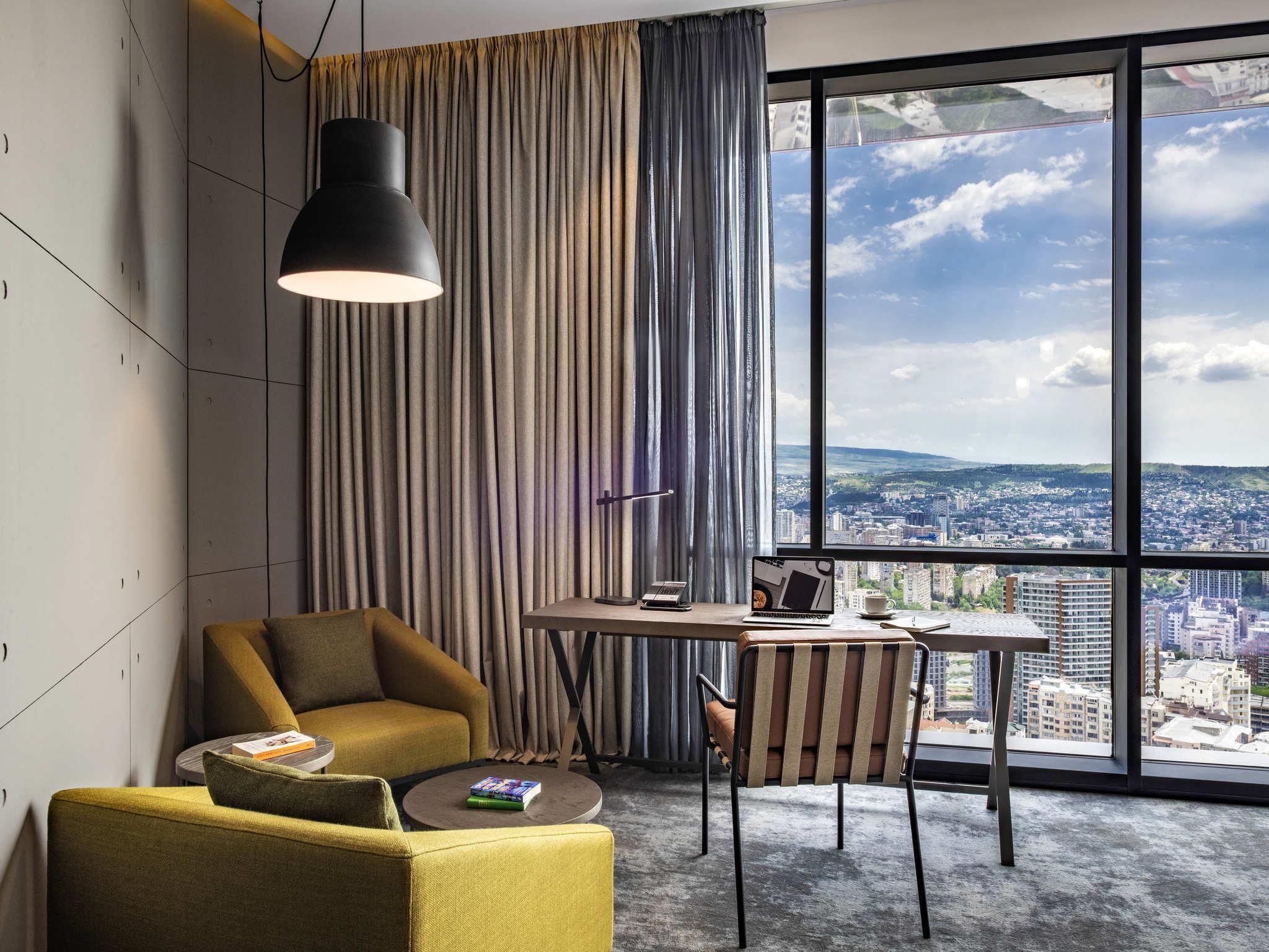 A modern hotel room with a large window, a desk, a chair, and a sofa.
