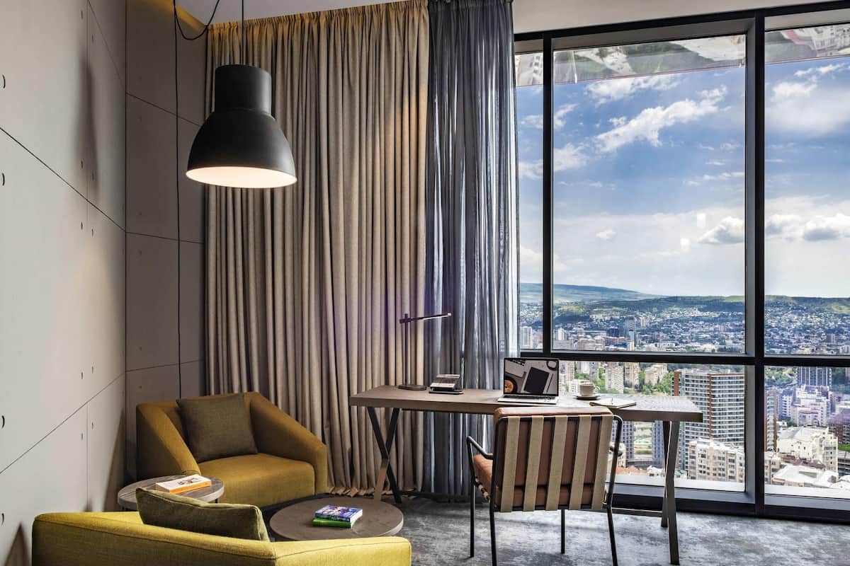 A modern hotel room with a large window, a desk, a chair, and a sofa.