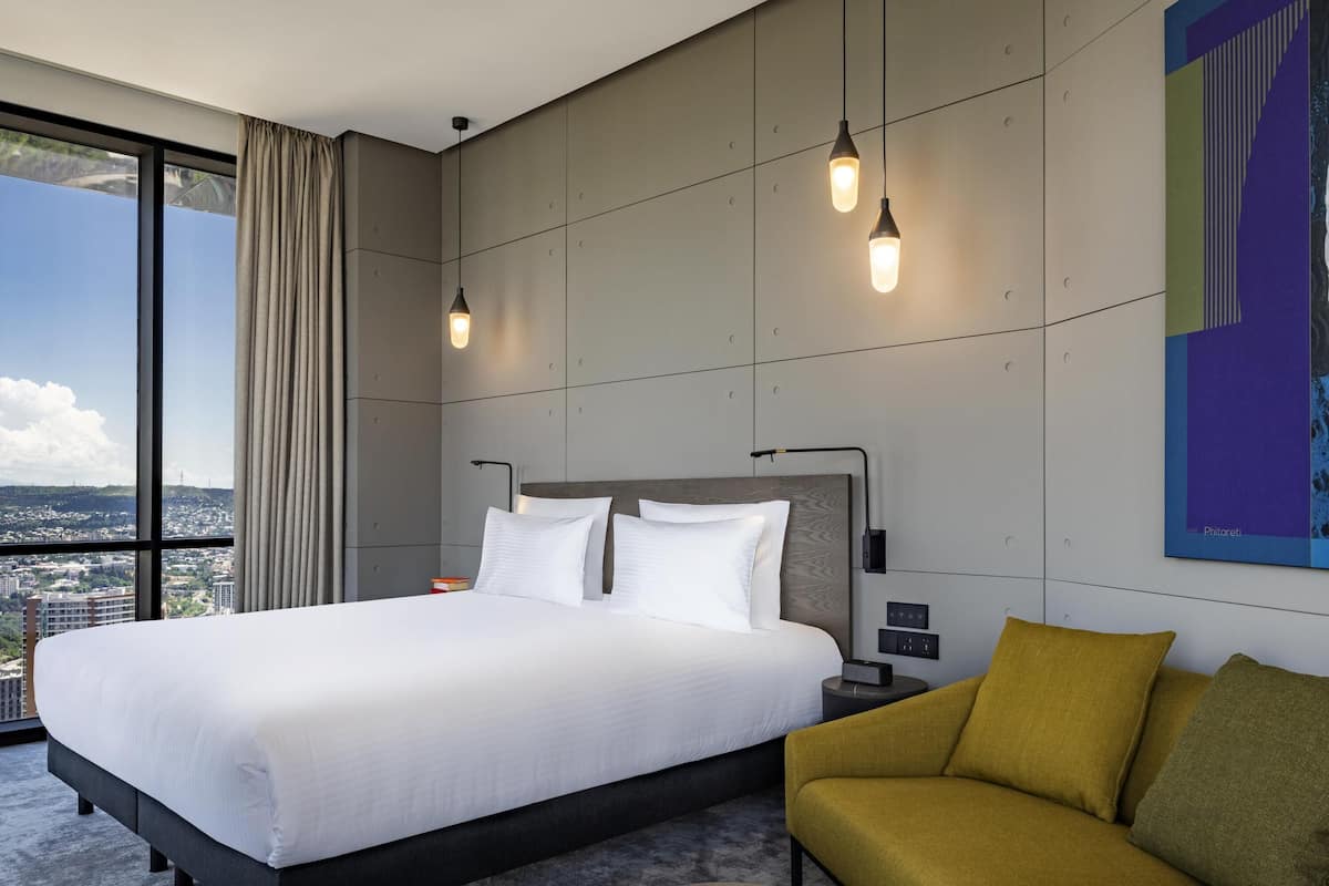 A modern hotel room with a large bed, a sofa, a round coffee table, and a city view.