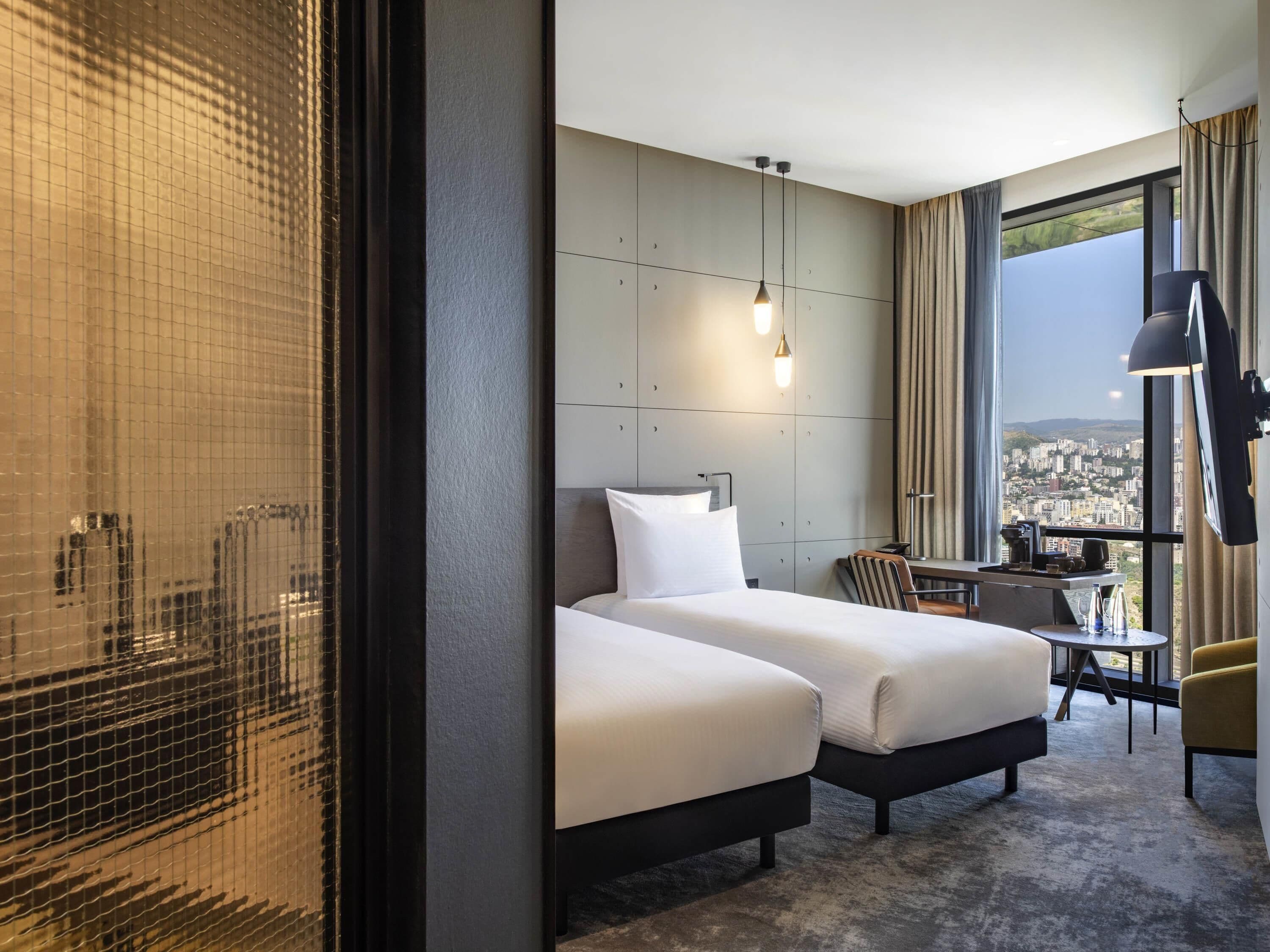 A hotel room with two beds, a desk, a chair, a large window with a city view, and a hanging light fixture.