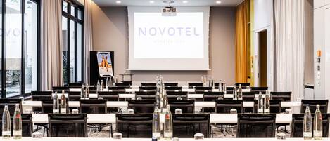 Un salón de conferencias con mesas, sillas y una pantalla de proyector que muestra 'NOVOTEL MUNSTER CITY'.