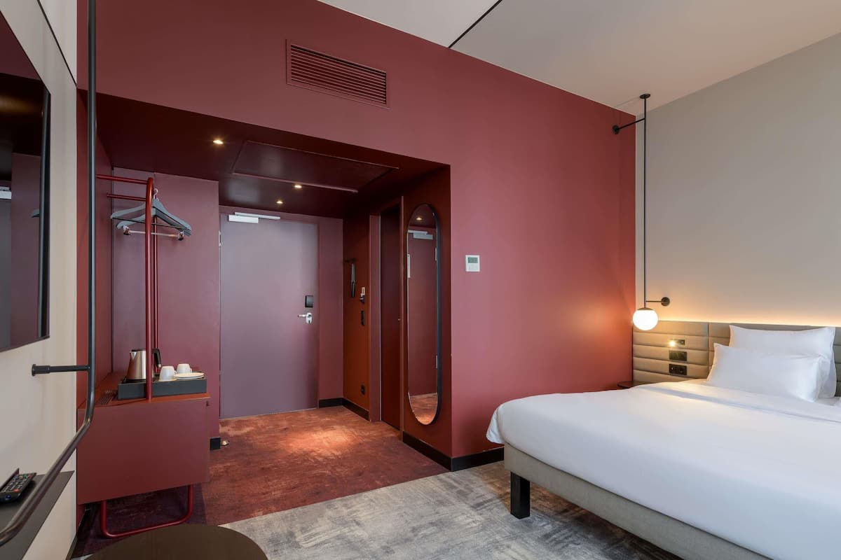 Una camera d'albergo con un letto, una scrivania, una sedia, uno specchio, un appendiabiti e una porta che dà su un corridoio.