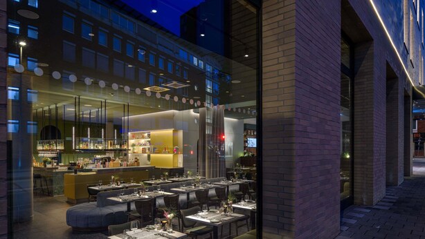 Moderni ravintola, jossa on baarialue, useita ruokapöytiä ja avoin keittiö.