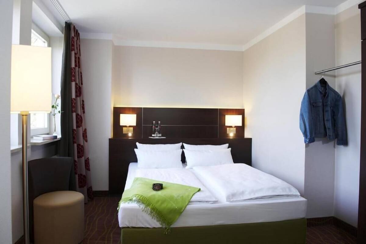 Habitación de hotel con cama, dos lámparas de noche, una silla, una chaqueta colgada y una ventana con cortinas.