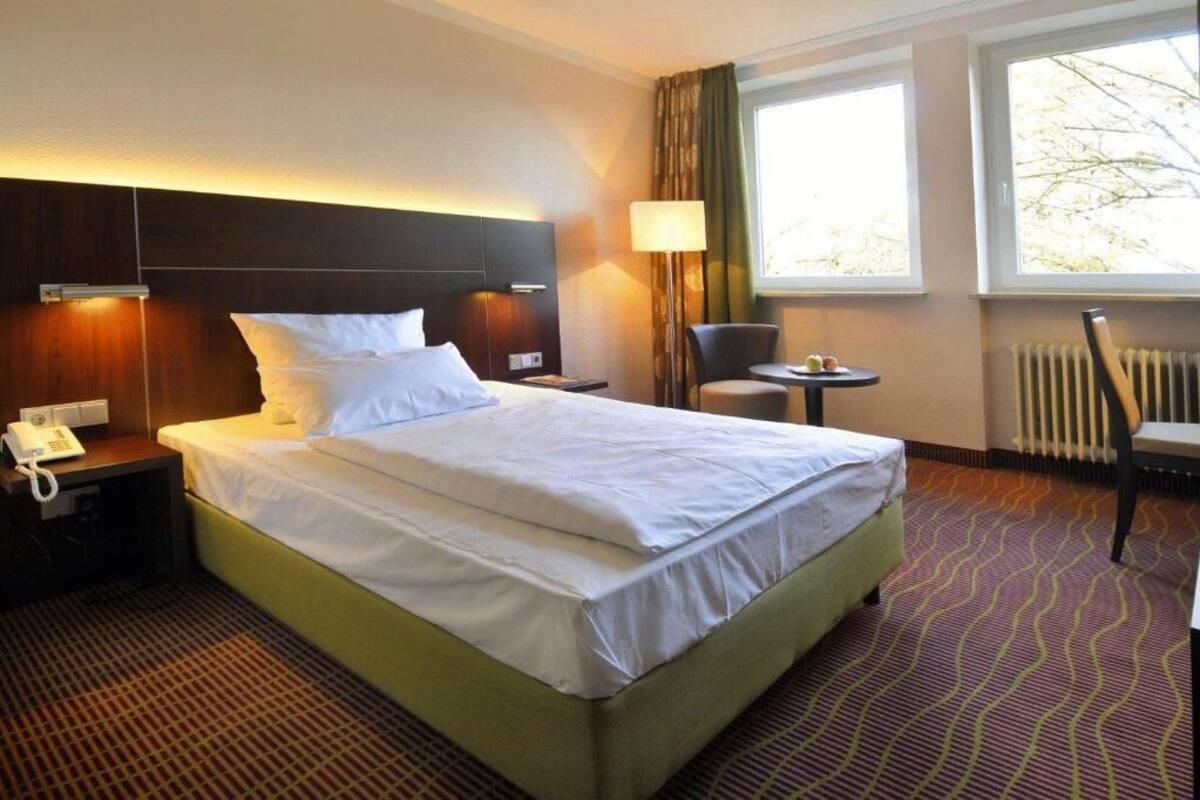 Habitación de hotel con una cama grande, una mesita de noche con lámpara, una silla y una ventana con cortinas.