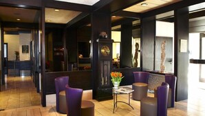A lobby with a reception desk, purple seating, a clock, and a vase with flowers. - Stern Hotel Soller (Ismaning)