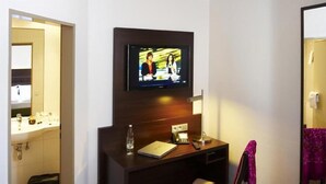 A hotel room with a TV mounted on the wall, a desk with a laptop, a bottle of water, and a lamp. - Stern Hotel Soller (Ismaning)