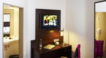 Habitación de hotel con televisión montada en la pared, escritorio con una computadora portátil, una botella de agua y una lámpara.