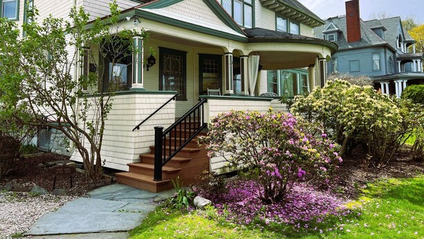 A two-story house with a porch, a staircase, and a garden with flowering plants. - Victorian Mansion Jim-Dandy Suite (Bennington)
