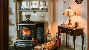 A room with a vintage stove, a mantelpiece with framed pictures, a table with a lamp, and a fire extinguisher. - Victorian Mansion Jim-Dandy Suite (Bennington)