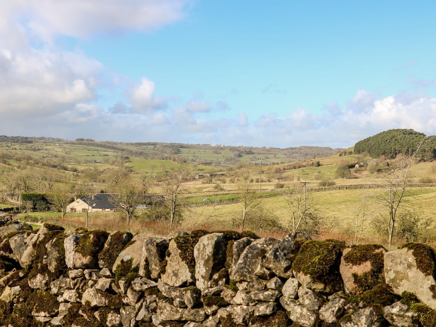 A stone wall in the foreground with a rural landscape in the background, including fields, a hill, and a small building.