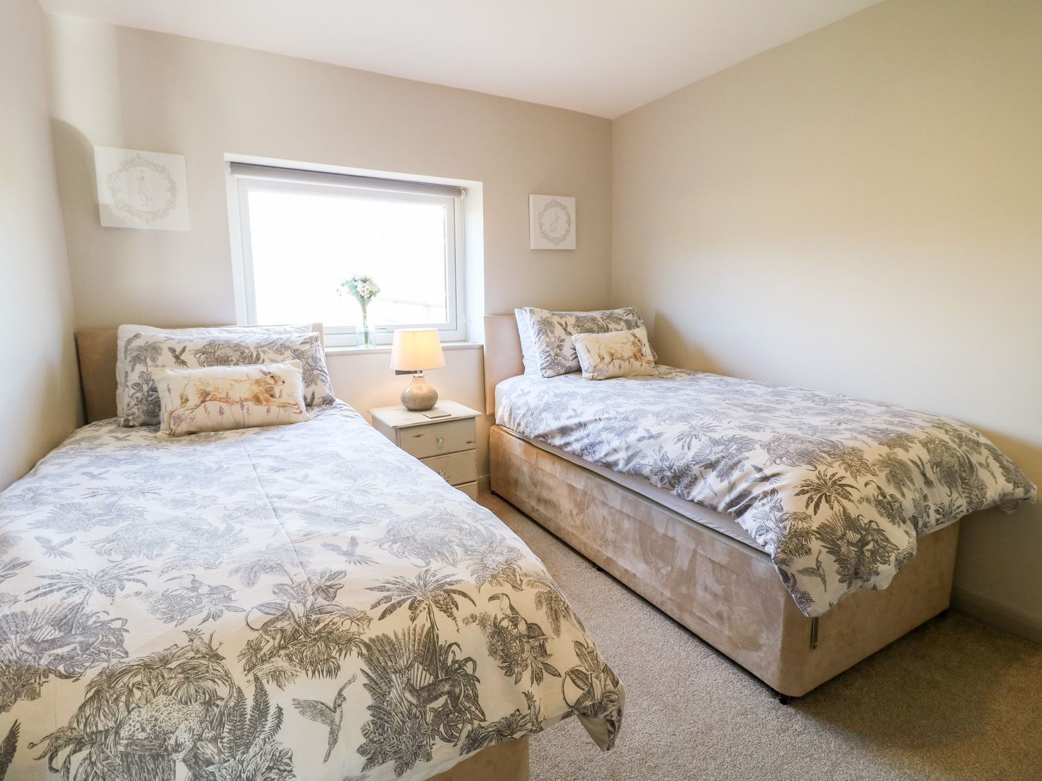 Une chambre avec deux lits, chacun agrémenté de draps à motifs floraux, une table de chevet avec une lampe et une fenêtre donnant sur un paysage verdoyant.