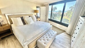 Ein Schlafzimmer mit einem großen Bett, einem Nachttisch, einer Lampe, einem Fenster mit Blick und einer Kommode.