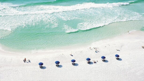 Ein Strand mit klarem türkisfarbenem Wasser und weißem Sand. Mehrere blaue Sonnenschirme und Liegestühle sind vorhanden.