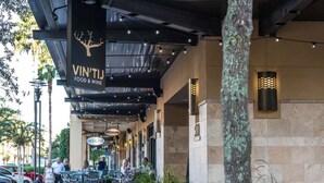 Ein Außenbereich mit Essbereich, Tischen, Stühlen und einem hängenden Schild für 'VIN'TIJ FOOD & WINE'. Im Vordergrund eine Mauer aus Stein und ein Baum.