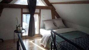 Ein Schlafzimmer mit schrägen Wänden, einem Bett, einem kleinen Tisch mit Pflanze und einem Fenster mit Vorhängen.