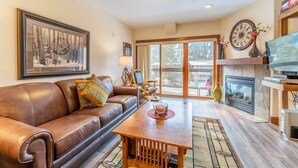 A living room with a brown leather sofa, a wooden coffee table, a fireplace, and a large window with a view of trees. - Private Balcony with Mountain Views; Close to Base Area & River Run Village! (Keystone)