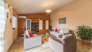 A modern living room with two sofas, a coffee table, and a TV. There is a kitchen area in the background with cabinets and a window. - Six Bedroom Villa with Pool (Kaštel Stari)