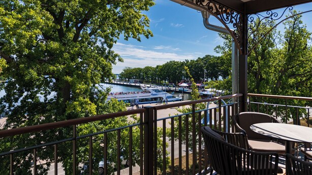 A balcony with a view of a docked boat, trees, and a clear sky. - Hafenhotel zu Putbus (Putbus)