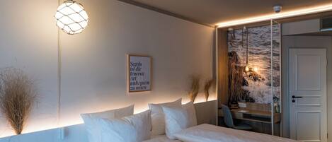 Quarto com cama, luminária na parede, frase emoldurada e vista do mar através de uma porta de vidro.