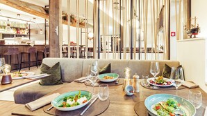 A dining area with a wooden table set for two, a bar in the background, and a view through a rope curtain. - Hafenhotel zu Putbus (Putbus)