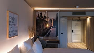 Un dormitorio con cama, escritorio con computadora, un cuadro en la pared y una puerta que da a otra habitación.