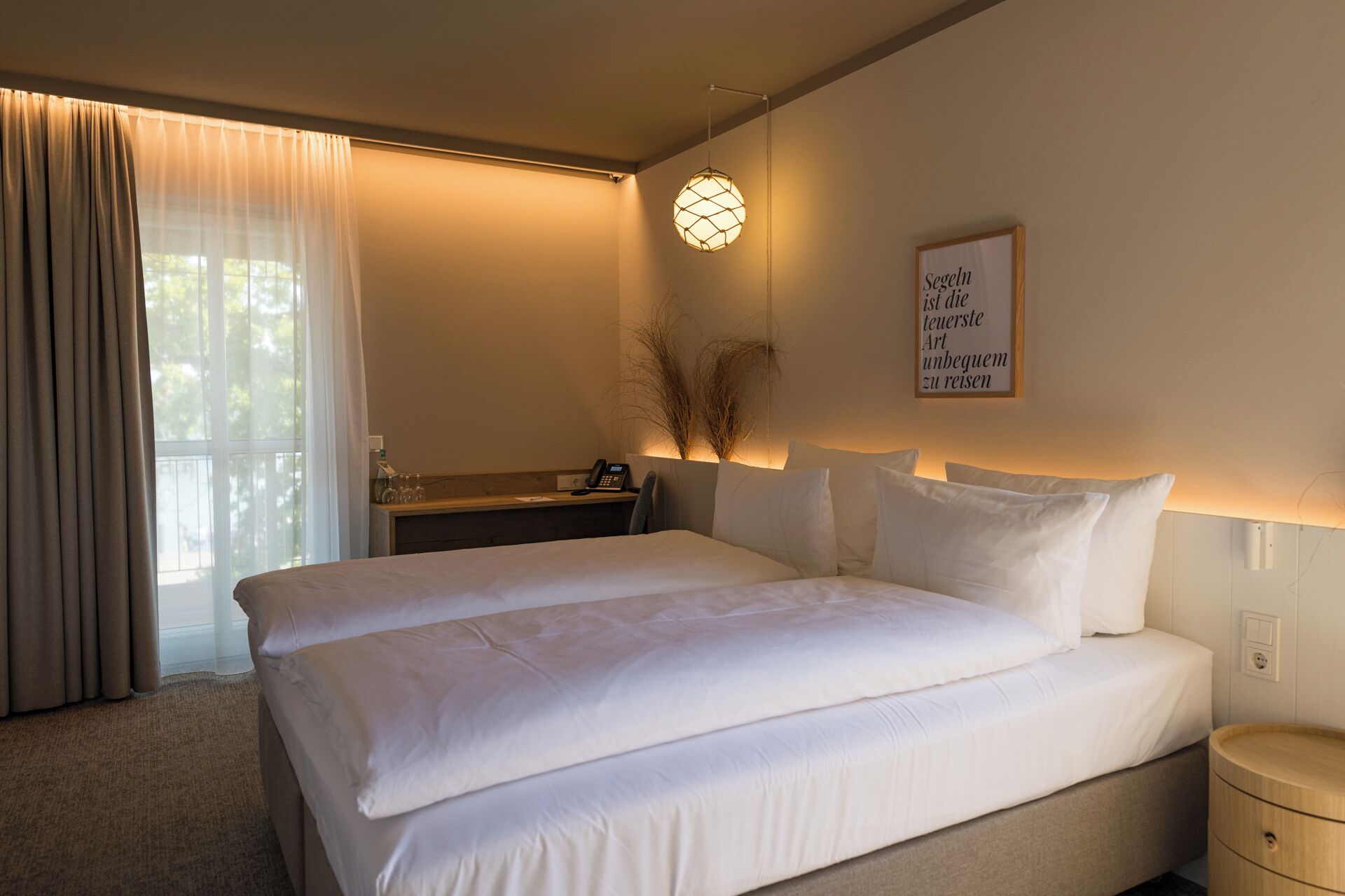 Een hotelkamer met een groot bed, nachtkastje, lamp, een raam met gordijnen en een citaat in een lijst op de muur.